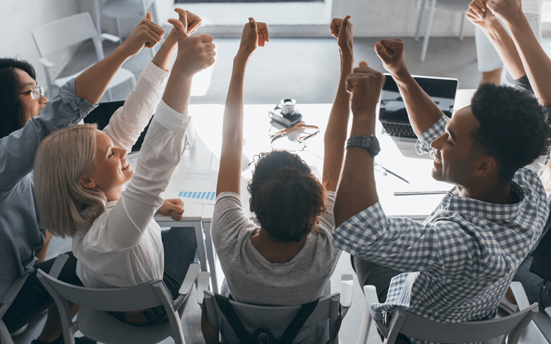 A imagem mostra um grupo de pessoas sentadas em uma mesa redonda no trabalho. A ideia é representar, de forma lúdica, a ausência de uma hierarquia rígida no modelo de gestão horizontal.