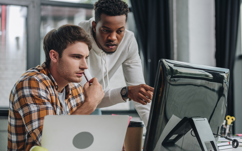 Dois jovens em roupas casuais conversando em frente a uma mesa com computadores. A imagem representa o RH em busca da cultura de inovação.