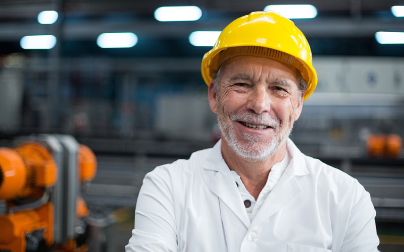 Na imagem vemos um senhor de barba branca e camisa social despojada utilizando capacete amarelo. Ele está em ambiente de trabalho e sorrindo, o que representa a motivação para trabalhar e como é importante motivar funcionários.
