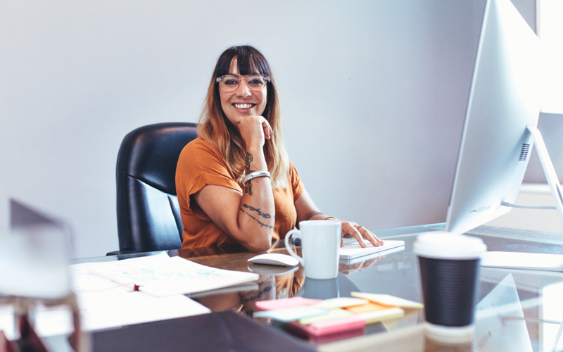 Na imagem, vemos uma jovem sorrindo sentada em uma mesa com papéis. Ela busca representar o papel do RH na reestruturação dos negócios.