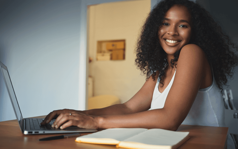 A imagem mostra uma mulher jovem sorrindo enquanto utiliza o notebook. Ela busca representar características comportamentais importantes para os profissionais.