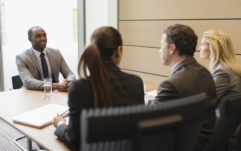 A imagem mostra quatro profissionais conversando. Um deles, do lado oposto da mesa, parece ocupar uma alta posição dentro da empresa. A imagem busca representar a importância de seguir um guia completo para um recrutamento preditivo eficaz.