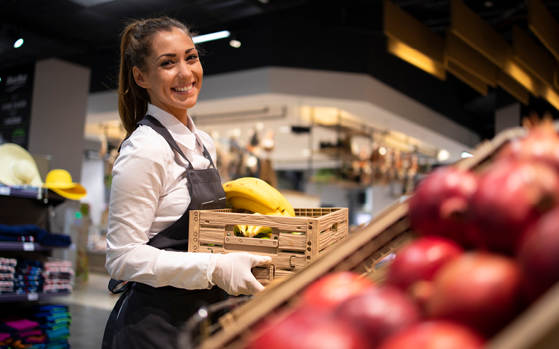 Mulher uniformizada trabalhando na reposição de frutas em um supermercado. A imagem representa uma funcionária temporária.
