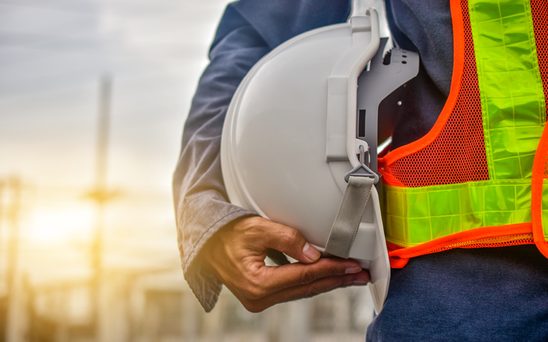 Lateral do corpo de homem utilizando colete e com capacete em mãos. A imagem representa trabalhadores temporários.