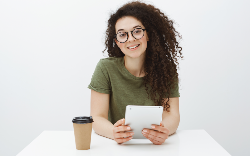 A imagem mostra uma jovem sentada em frente a uma mesa segurando um tablet e com um copo de café ao lado. A imagem busca representar a importância de qualificar candidatos em processos seletivos.