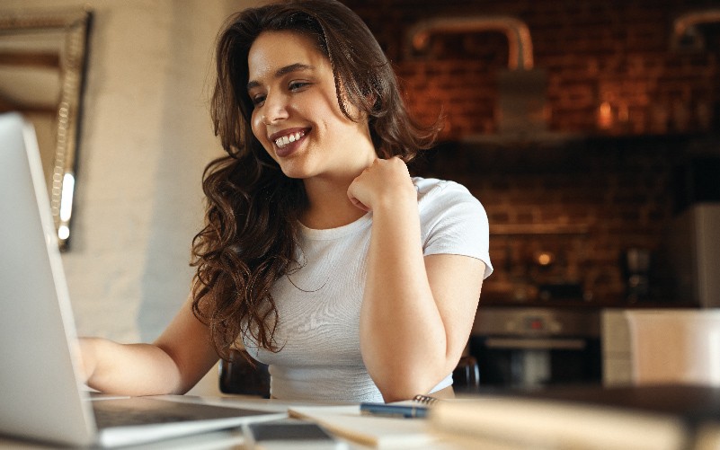 A imagem mostra uma mulher sentada em uma mesa. Ela está mexendo no notebook e sorrindo, feliz ao observar os bons resultados obtidos por meio da análise de indicadores de recrutamento e seleção da empresa que ela trabalha como analista de RH.