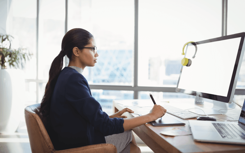 Na imagem, vemos uma mulher sentada olhando para a tela do computador enquanto faz anotações. A imagem busca representar a importância do protagonismo do RH para o sucesso das organizações.