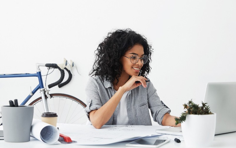 A imagem mostra uma jovem, de cabelos cacheados, sentada em uma mesa estudando sobre a diferença entre programa de trainee e programa de estágio em um notebook.