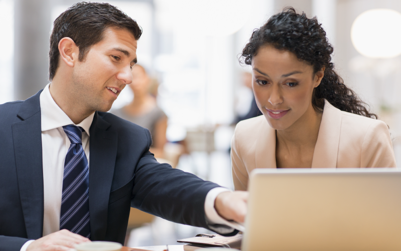 A imagem mostra dois profissionais, um homem e uma mulher, conversando enquanto observam algo no computador. A imagem busca representar, de forma lúdica, como atua o headhunter e como são feitos os recrutamentos de alta complexidade.