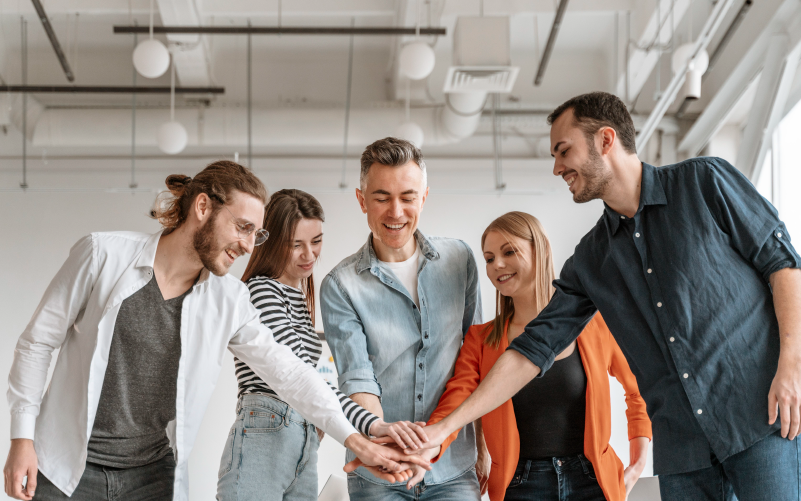 Na imagem, vemos um grupo de pessoas sorrindo, com a mãos unidas ao centro. A imagem busca representar, de forma lúdica, como deve ser a experiência dos funcionários na sua empresa, ou seja, a employee experience.