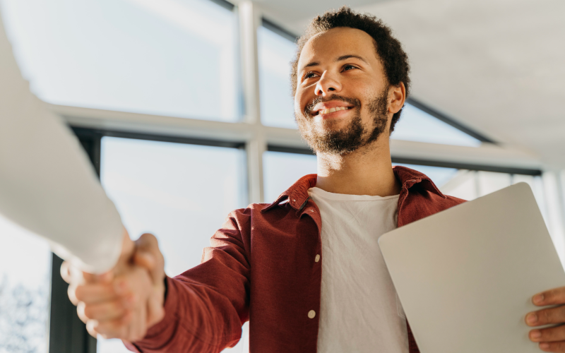 A imagem mostra um homem sorrindo enquanto realiza um aperto de mãos com alguém. Ele segura seu currículo e representa os benefícios do outsourcing no recrutamento e seleção.