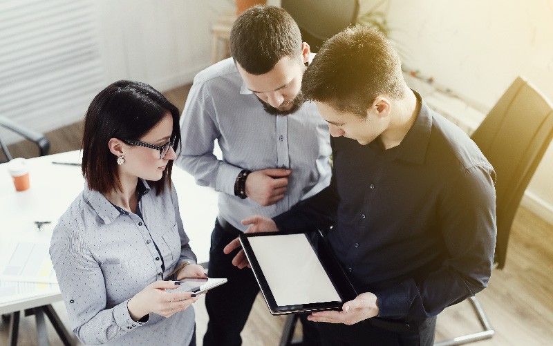 A imagem mostra três profissionais conversando enquanto olham para a tela de um tablet. Ela busca representar, de forma lúdica, a análise de soft skills de candidatos e colaboradores.