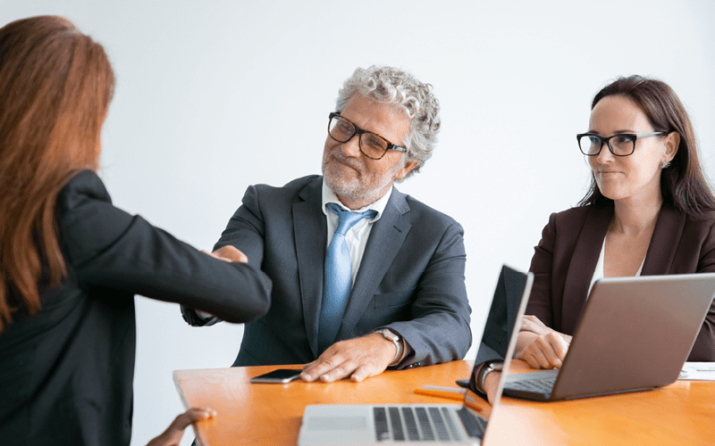 A imagem mostra duas jovens conversando com um senhor de cabelos grisalhos. As jovens estão com notebooks e uma delas está realizando um aperto de mão com o senhor. A imagem busca representar como a inovação tecnológica já faz parte da rotina do RH das empresas.