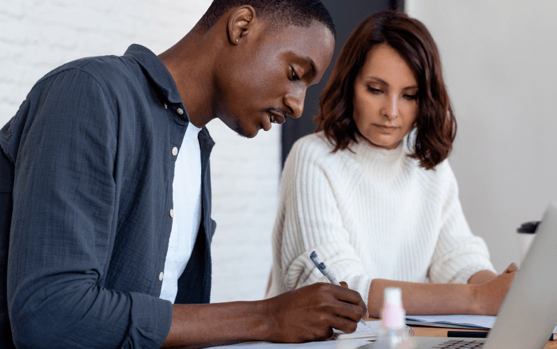 Na imagem vemos um homem e uma mulher sentados lado a lado. O homem parece estar assinando documentos de contratação para trabalho temporário.