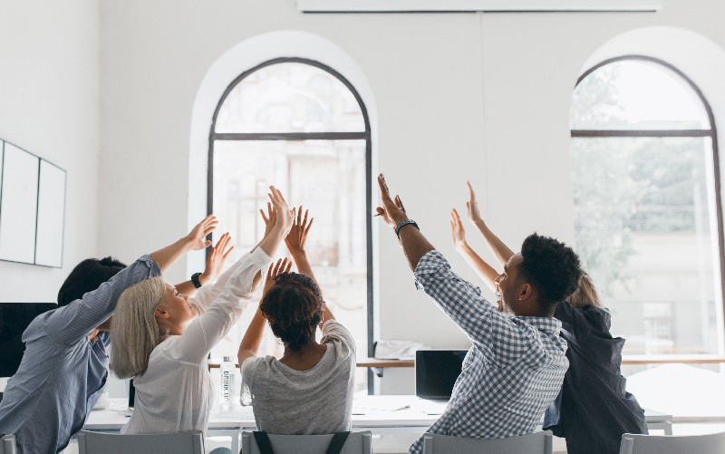 A imagem faz referência a uma cultura organizacional positiva ao mostrar um grupo de colaboradores celebrando juntos. Todos estão sentados ao redor de uma mesa, com os braços erguidos.