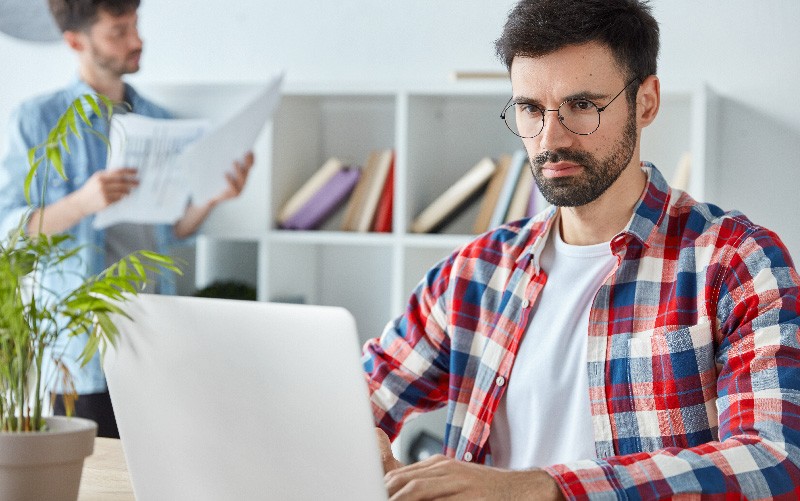 A imagem mostra um homem jovem e branco fazendo pesquisas sobre RH e Lei Geral de Proteção de Dados em um notebook. Este homem é jovem, branco, usa óculos e tem barba.