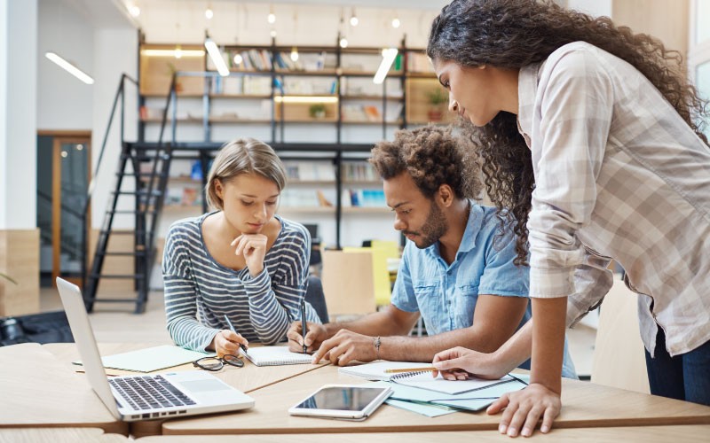 Na imagem, vemos três jovens conversando ao redor de uma mesa enquanto observam papéis. A imagem busca representar como funciona a consultoria para contratação em cargos de gestão.