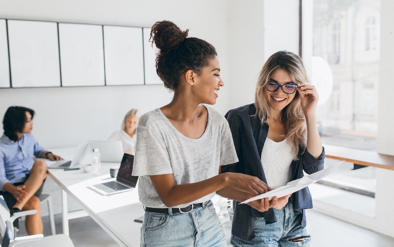 Na imagem vemos duas jovens, uma de cabelos cacheados presos em um coque e outra de cabelo loiro e solto. Elas conversam felizes enquanto caminham por uma sala. A imagem busca representar como uma avaliação de desempenho bem sucedida contribui para um bom clima organizacional e para a conquista de bons resultados.