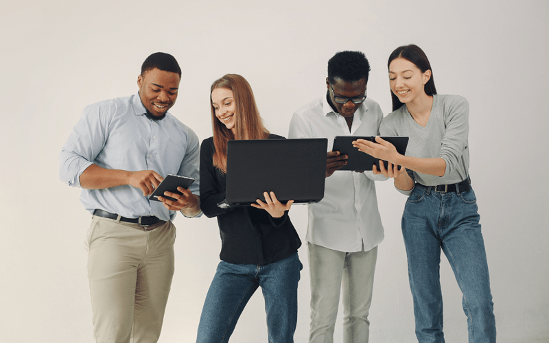 A imagem mostra dois homens negros e duas mulheres brancas conversando sobre processos de estágio e trainee. Eles estão animados e mostram informações de notebooks e tablets uns para os outros.