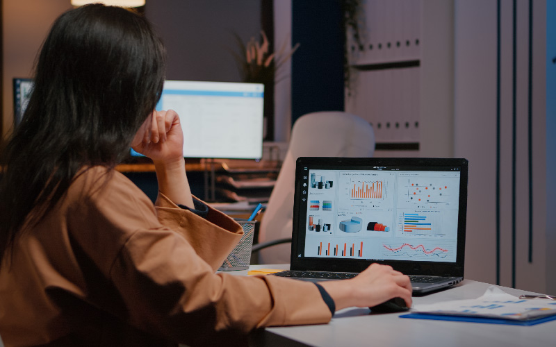 A imagem mostra uma mulher sentada em frente ao computador, trabalhando. Ela está de costas e o foco da imagem são os gráficos em sua tela. Ao fundo, vemos mesas e computadores, como em um escritório. A imagem busca representar as mudanças para gente e gestão digital, se adaptando para o RH tecnológico.