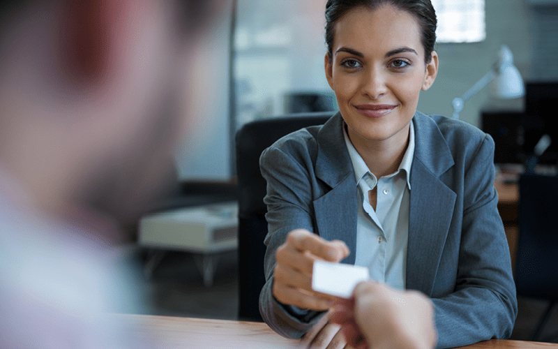 Na imagem, vemos uma mulher em roupas sociais entregando um papel para um homem, que está em desfoque. A imagem mostra a importância de saber recrutar desenvolvedores de software.