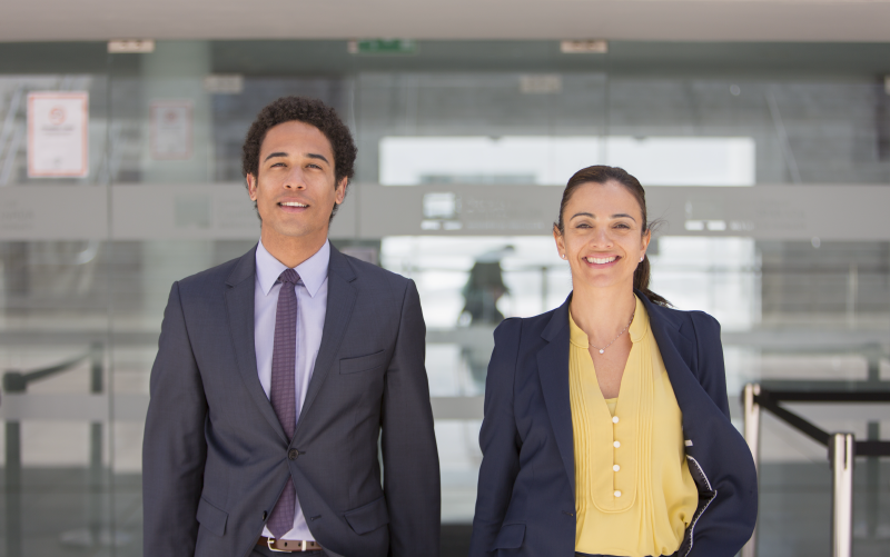 Na imagem, vemos um homem e uma mulher lado a lado, ambos de pé e com trajes sociais. Aparentemente, eles estão em um ambiente corporativo. A imagem busca representar a importância da equidade de gênero nas organizações.