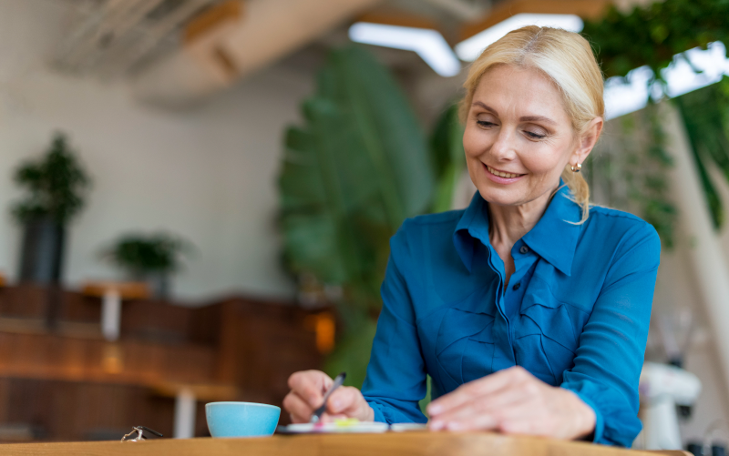 Na imagem, vemos uma mulher sentada em frente a uma mesa e fazendo anotações. Ela é aparentemente mais velha e busca representar o preconceito etário, ou etarismo, nas empresas.