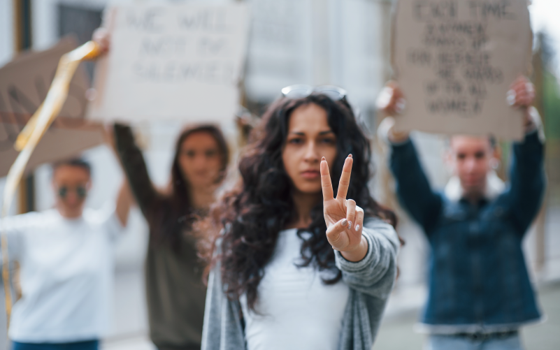 Na imagem, vemos uma mulher em destaque. Ela está com semblante sério e faz o gesto de "paz" com as mãos (dois dedos erguidos). Ao fundo, em desfoque, está um grupo de pessoas levantando cartazes. A imagem busca representar a importância dos movimentos sociais e como as empresas se apropriam deles fazendo causewashing para lucrar.