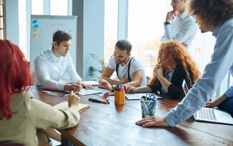 A imagem mostra jovens sentados ao redor de uma mesa conversando e analisando papéis. A imagem busca representar, de forma lúdica, a análise e o planejamento de como fazer programa de estágio na empresa que trabalham.