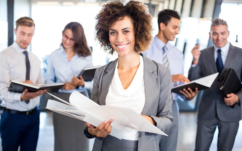 Na imagem, vemos uma mulher em destaque, segurando papéis e liderando uma equipe, que está ao fundo. Ela mostra como liderar bem um equipe.