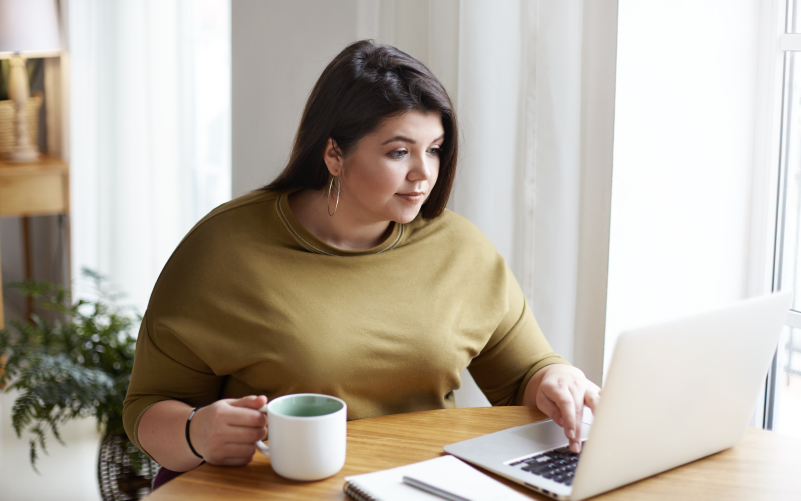 A imagem mostra uma mulher tomando café e trabalhando em um notebook. Ela está mais séria, pois está atuando no processo de hunting no RH na empresa onde trabalha. Ao fundo, observa-se uma sala clara, iluminada e com móveis em madeira.