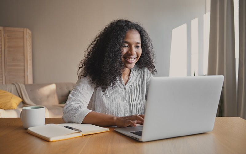 A imagem mostra uma mulher negra trabalhando em um notebook. Ela está sorrindo, enquanto entende melhor sobre o engajamento dos colaboradores na empresa onde trabalha.