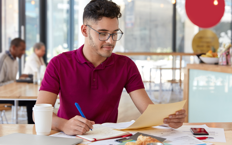 A imagem mostra um jovem branco estudando sobre os principais testes de recrutamento para seleção de pessoal. Ele está sentado em uma mesa, com diversos papeis e anotações a seu redor. Ele usa óculos, tem barba e usa uma camisa vermelha.