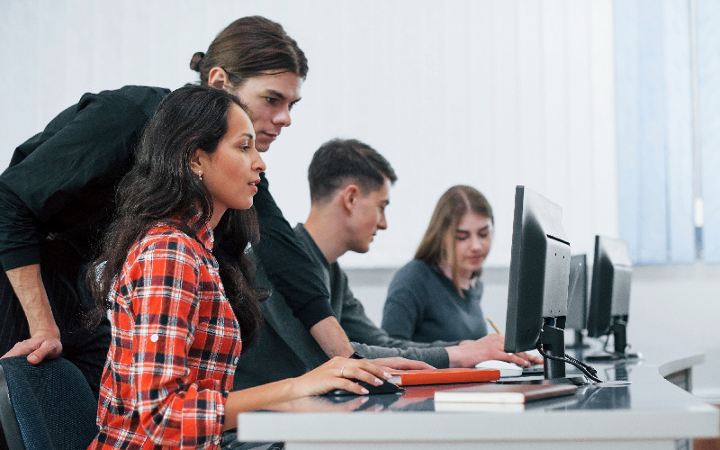 A imagem mostra alguns jovens sentados em uma mesa, cada um em frente a um computador. Em destaque, vemos uma mulher concentrada na tela do computador, sendo auxiliada por um homem, que está em pé e utilizando o mouse. A imagem busca representar de forma lúdica os conhecimentos em tecnologia e a importância de contratar profissionais de TI.