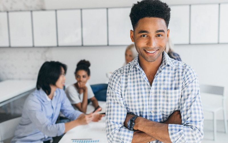 A imagem mostra um homem jovem e negro em foco. Ao fundo, observa-se mulheres conversando sobre como fazer gestão de pessoas e boas práticas para isso.