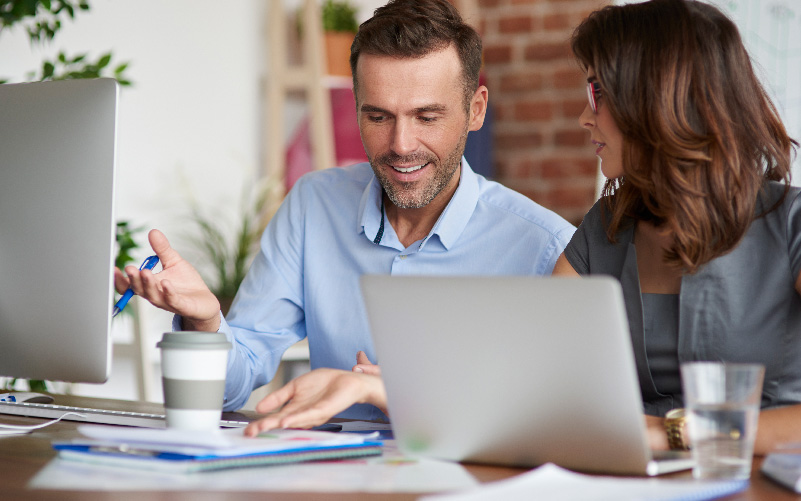 A imagem mostra um homem e uma mulher trabalhando em um projeto de consultoria de RH. Eles estão conversando, mexendo em papeis e analisando relatórios.