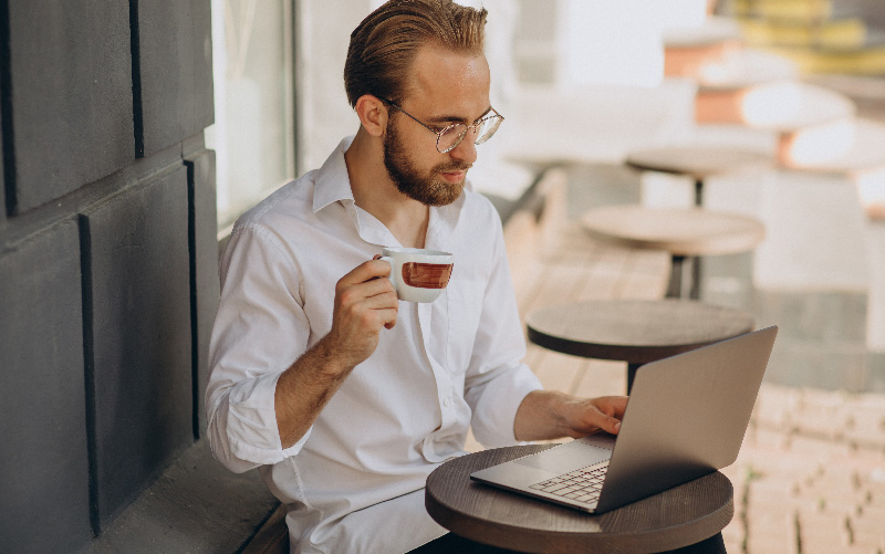 A imagem mostra um homem branco tomando café e pesquisando em um notebook sobre o que é e como funciona uma plataforma de recrutamento e seleção. Ele é jovem, está vestindo uma camisa social branca e usa óculos.