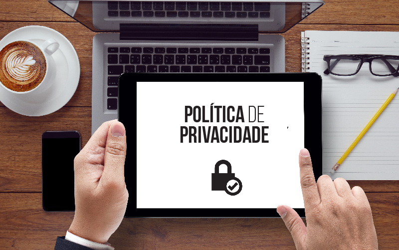 A imagem é focado nas mãos de um home segurança um tablet. No dispositivo, está escrito política de privacidade em destaque. Ao fundo da imagem, é possível observar também um notebook.