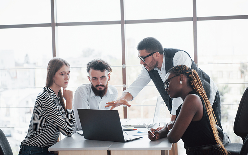 A imagem mostra um grupo de quatro pessoas diversas conversando sobre a relação entre desenvolvimento profissional e propósito, analisando informações e dados sobre pessoas em um notebook.