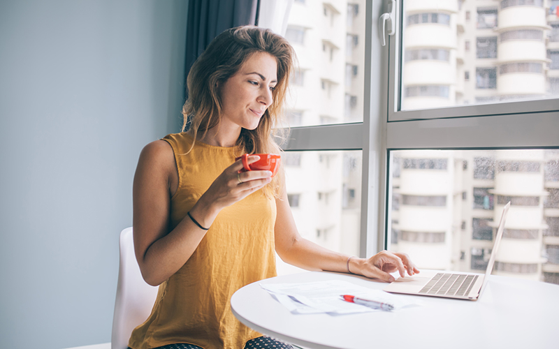 A imagem mostra uma mulher branca e loira como foco. Ela está tomando café enquanto pesquisa em um notebook sobre a aplicação da psicologia positiva na gestão de pessoas.