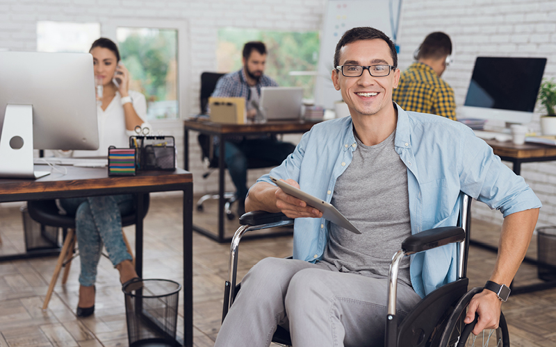 A imagem mostra um home de cadeira de rodas em um escritório segurando um tablet ao lado de seus colegas de trabalho. Ele está feliz por ter encontrado seu trabalho com propósito.