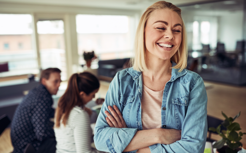 Mulher sorrindo por ter passado por um processo de job rotation na empresa que trabalha como trainee. Ela é jovem, loira e branca, e está vestindo uma camisa branca e jaqueta jeams. Está em pé em uma sala onde ao fundo observamos outras duas pessoas jovens trabalhando.