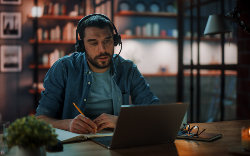 Jovem atuando no modelo de trabalho híbrido. Ele está participando de uma reunião online e fazendo anotações em um bloco de papel.