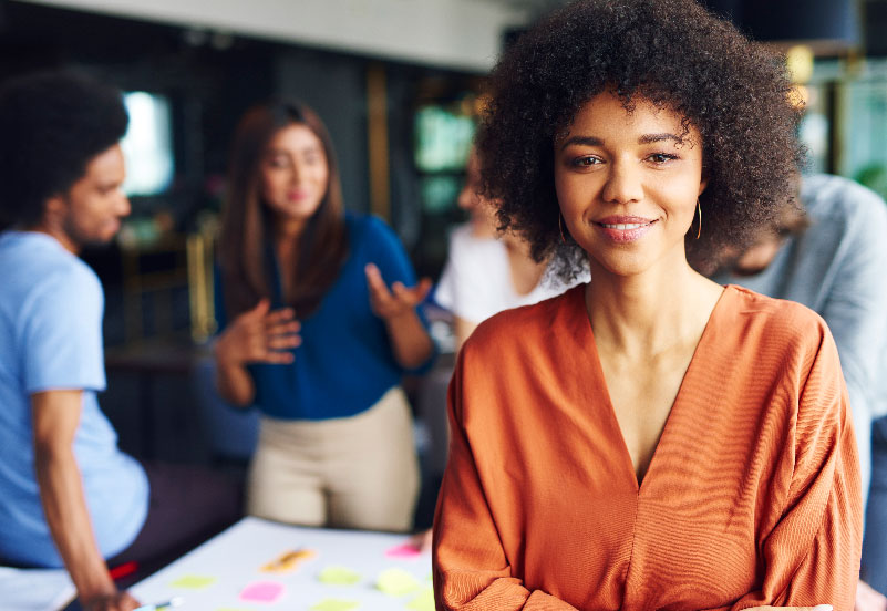 Imagem em ambiente de escritório, com pessoas ao fundo. Na frente, em destaque, mulher negra com roupas coloridas. Ela está com os braços cruzados e sorrindo, buscando representar de forma lúdica a figura da liderança e os modelos de gestão de pessoas.