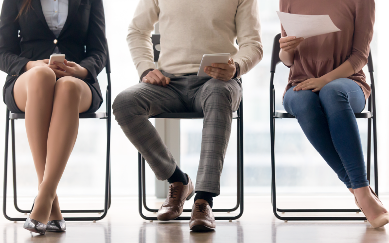 Três pessoas sentadas lado a lado, como em um processo seletivo. A imagem, que está recortada na altura da cintura das pessoas, dando foco à ação de estar sentado e às pernas, busca representar de forma lúdica as etapas de Business Case e Challenge Lab na seleção.