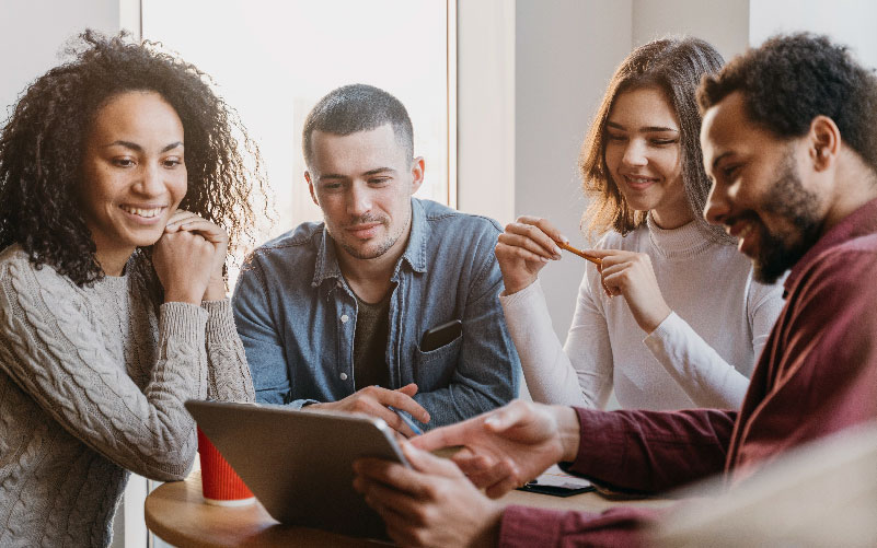 Imagem de quatro jovens profissionais sentados ao redor de uma mesa, conversando animados enquanto olham informações em uma tela. O objetivo é representar o planejamento de políticas de ESG nas empresas.