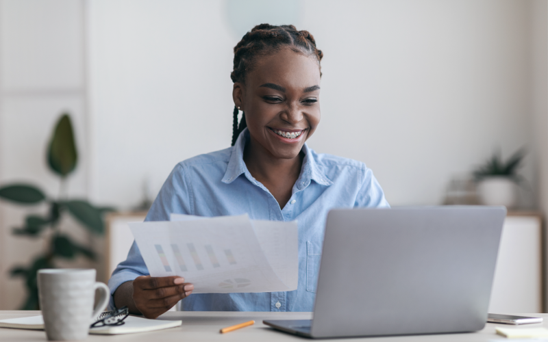 Imagem de jovem sorrindo em frente ao comutador, enquanto segura folhas de papel com uma das mãos. Ela busca representar o lifelong learning na prática.