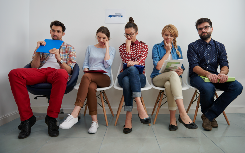 Imagem com cinco pessoas sentadas lado a lado, como em uma sala de espera. A imagem busca representar candidatos participando de um processo seletivo com recrutamento misto.