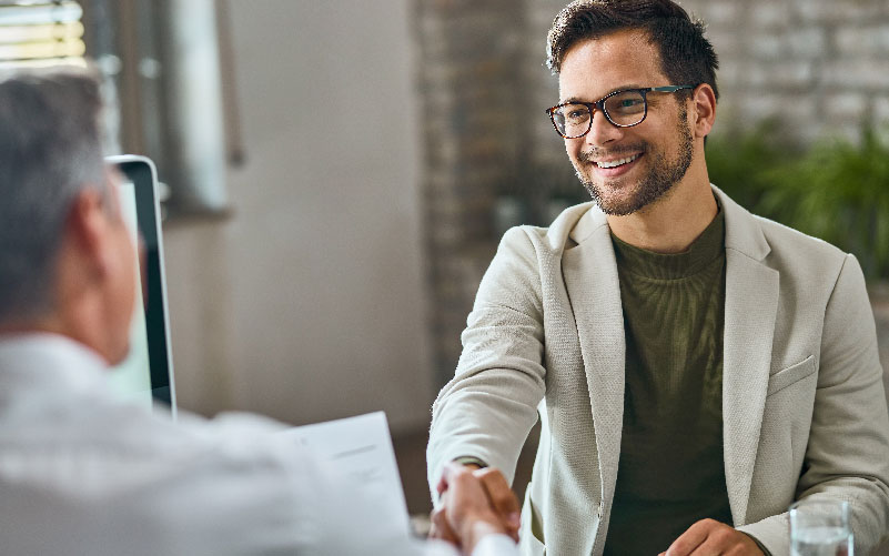 Imagem de duas pessoas realizando um aperto de mão e sorrindo. O objetivo é representar uma contratação nos diferentes tipos de recrutamento.