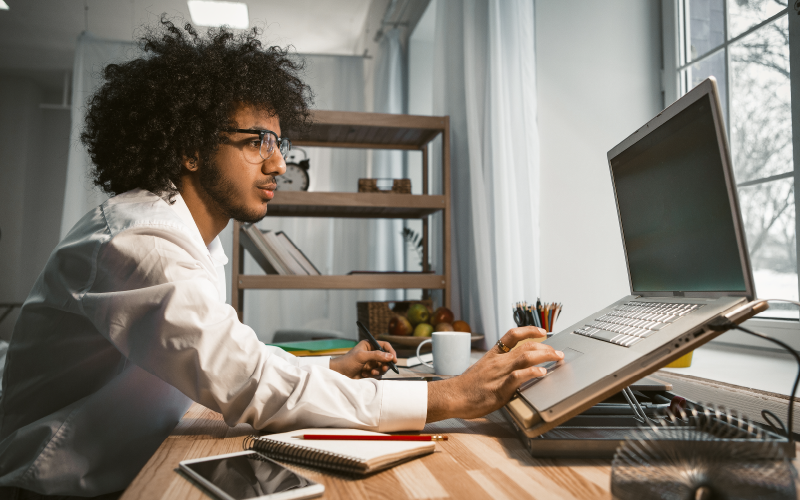 Imagem lateral de homem negro de cabelos cacheados e utilizando roupa social digitando algo em computador, que está a sua frente. Ele parece estar trabalhando em home office e, com isso, a imagem busca mostrar os novos desafios do profissional de RH.